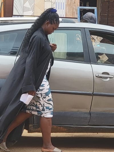 This daytime image, set in Maiburiji, Nigeria, captures a woman resting by a light-colored car in an outdoor setting. The woman, with dark skin and long, dark braided hair secured by a blue tie, is the central figure. She wears a long, dark robe or coat over a white garment with a distinctive blue and green camouflage-like pattern. She is holding a folded white piece of paper in her right hand, and a dark smartphone is tucked into her waistline on her right side. Her expression is neutral as she looks downwards and to her left. Her right leg is bare, revealing a bare foot, while her left foot wears a light-colored sandal.
She is positioned against or leaning on a silver or light grey four-door car, which appears to be a van or SUV. A car door handle is visible on the rear passenger door. In the reflection of the driver's side mirror, the head of another person wearing a dark cap can be seen.
The background reveals parts of buildings or structures with varied tiled exteriors, including white, grey, and patterned tiles, along with some exposed brickwork. The ground is a light brown, earthy color. The bright lighting suggests clear weather conditions. No specific text is clearly visible in the image. The scene conveys a sense of a casual break or waiting moment in what appears to be an urban or semi-urban environment.