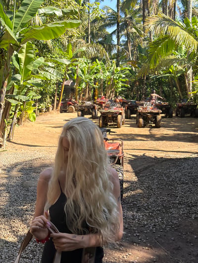 The image displays an outdoor scene in Payangan, Indonesia, centered around a gathering of all-terrain vehicles (ATVs) amidst lush tropical foliage. In the immediate foreground, positioned slightly to the left, a woman with long, wavy blonde hair cascading over her face is seen from the chest up. She wears a black spaghetti-strap top and her head is angled downwards, obscuring her features. Her hands are clasped at waist level, holding a light-colored strap or object, with a red bracelet visible on her left wrist.
The background is dominated by dense, vibrant green vegetation, including numerous large-leafed banana trees and tall palm trees, suggesting a tropical or jungle environment. A dirt path, featuring both compacted earth and gravel sections, stretches into the mid-ground, leading to a clearing. In this clearing, approximately ten red and black ATVs are parked in rows. One individual, wearing a dark helmet and clothing, is visible mounted on an ATV in the middle-right section of the background. Strong, direct sunlight illuminates the scene, casting distinct shadows on the ground. A small, rustic wooden structure is partially visible among the trees on the left side of the mid-ground, with some terracotta pots nearby.