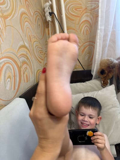 In this indoor shot, a young boy is lying on a couch, holding a smartphone. He is smiling broadly, looking up and to his right, with his head resting on a light-colored pillow. A stuffed fox toy is visible behind him on the couch. A person's foot, with red nail polish on the big toe, is in the foreground, obscuring part of the frame. The room appears to be a bedroom or living area, with patterned wallpaper and a white curtain. A power strip with a black cord is mounted on the wall in the background. The overall scene suggests a casual, relaxed moment at home.
