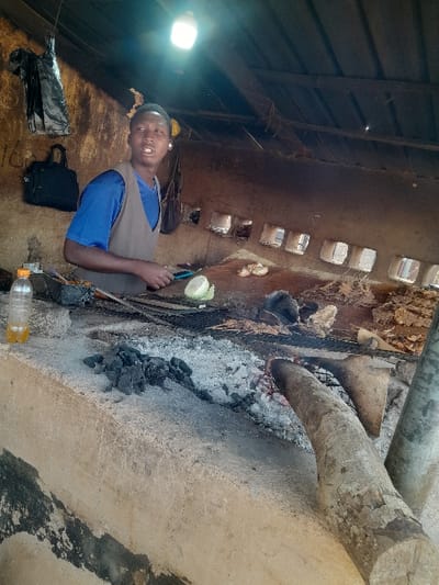 Within an unadorned, semi-enclosed structure in Kirana, Nigeria, a young male is depicted preparing food over an open fire. The structure features rough, unpainted walls, possibly mud or concrete, with a dark corrugated metal roof supported by wooden beams. A bare LED light bulb hangs from the ceiling, illuminating the scene. A horizontal band of small, rectangular openings is visible along the upper section of the back wall.
The male, positioned in the left-center, is wearing a blue t-shirt under a brown sleeveless vest. He holds a small, light-colored object (resembling a piece of cabbage) in his left hand and a cutting tool in his right hand, poised above a metal grill rack. His mouth is open, and he looks slightly to his right.
The cooking area is a substantial, rough concrete or mud block structure. On its surface, a metal grill holds multiple pieces of grilled meat, some appearing charred. A piece of green vegetable is also on the grill. To the left, a clear plastic bottle containing a yellowish liquid rests on the counter. Piles of charcoal and ash are visible on the cooking surface, with glowing embers and two large wooden logs serving as fuel sources to the right.
In the background, on the left wall, a black plastic bag hangs, and beneath it, a dark bag (possibly a satchel) is suspended. Faint markings, including the number "16," are visible on the wall. The overall environment appears functional and well-used, characteristic of a roadside food stall.
