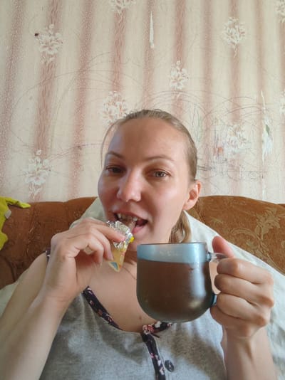 A young woman is sitting on a couch, holding a mug of dark liquid in her right hand and a wrapped candy bar in her left hand, with the candy bar partially in her mouth. She is looking directly at the camera. The background consists of floral wallpaper and the armrest of the couch. The scene appears to be an indoor setting, likely a home, in Чухлэм, Russia. The lighting is natural, coming from an unseen source.