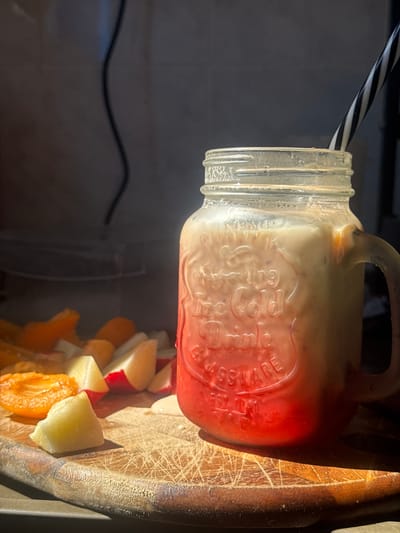 The image displays a clear glass mason jar mug containing a layered beverage, positioned on a well-worn wooden cutting board. The drink features a vibrant red layer at the bottom, transitioning into an opaque, light beige top layer with visible speckles. A black and white striped straw is inserted into the drink.

The glass mug is embossed with decorative text that includes phrases such as "Drinking," "Ice Cold," "Drink," and "GLASSWARE EST 1941."

To the left of the mug, several pieces of cut fruit are scattered on the cutting board. These include a halved orange-colored fruit with an exposed core, light yellow-green fruit chunks, and red-skinned fruit wedges with white interiors.

The wooden cutting board shows significant wear, marked by numerous knife cuts and scratches. The scene is illuminated by a strong, directional light source from the left, highlighting the subjects. The background is dark, out of focus, and indistinct, with a wavy black cable visible in the upper left quadrant. No individuals are present in the frame.