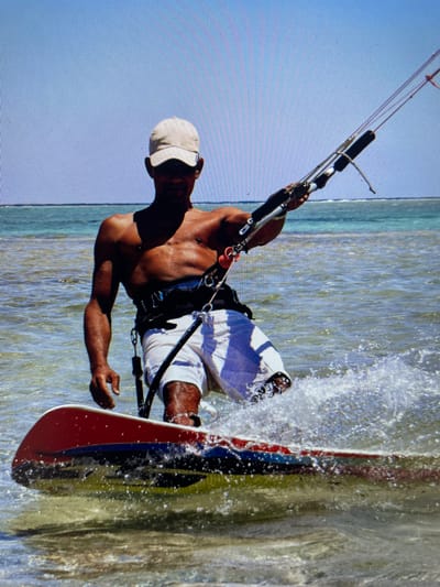 A man kitesurfs on water. He wears a white hat, dark harness and white and blue shorts. He's holding a kite with both hands. A red, blue and black board is partially submerged in water. The water is murky with whitecaps. The sky is light blue. The location is likely in Bacong, Philippines, based on the image's origin.