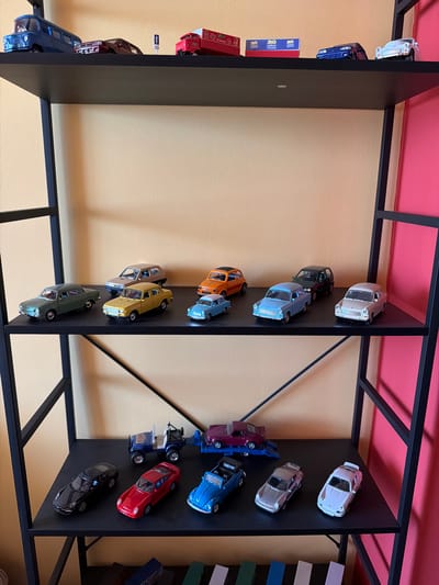The image shows a collection of model cars displayed on black shelves against a yellow wall. The shelves are part of a larger structure with black metal framing and a pink ceiling. The model cars are of various colors, including silver, blue, red, orange, yellow, green, and black. They are arranged in rows on different shelves. Some of the cars are vintage models, while others appear more modern. There are also some books or boxes with text on them placed on the shelves. The overall scene suggests a display or collection, possibly in a home, shop, or museum. There are no people visible in the image. The lighting appears to be artificial, suggesting an indoor setting. The image is oriented such that the shelves and cars are viewed from a high angle, looking downwards.