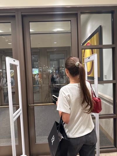 A young Caucasian woman with her back to the camera is standing in front of double glass doors. She has long brown hair tied in a ponytail with a scrunchie and is wearing a white t-shirt and dark jeans. She is holding a black bag in her left hand and a red purse on her right shoulder. In the foreground to the left is a white security gate. The glass doors reflect people and store displays. In the background on the right is a piece of abstract art with yellow and red colors. The scene appears to be inside a retail store in Coral Gables, Florida.