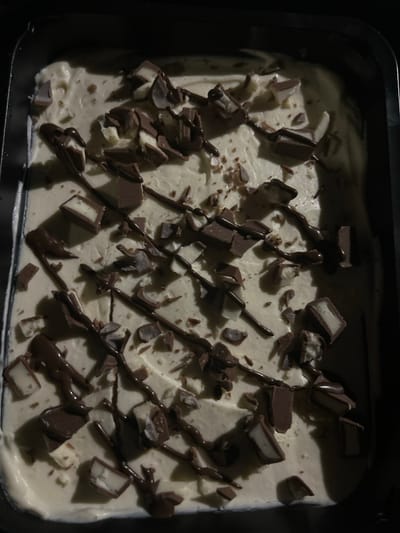 An overhead, close-up image displays a light-beige, creamy dessert within a dark, rectangular pan. The dessert surface is adorned with various chocolate toppings: a dark brown, glossy liquid drizzle in irregular diagonal and zig-zag patterns, numerous solid dark chocolate pieces of diverse sizes and shapes, and several dark chocolate segments distinctly revealing white or light-colored interiors. Additionally, smaller chocolate shards are scattered throughout. The scene is lit from the upper right, creating subtle shadows and highlighting surface textures. The image provides no discernible visual information to identify the location as Bischheim, France.