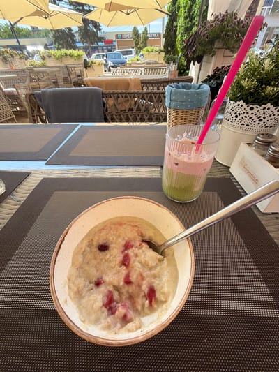 A close-up, eye-level shot captures a breakfast scene on an outdoor patio, likely in Leningradskaya, Russia. In the foreground, a bowl of oatmeal with visible red berry pieces sits on a dark brown placemat. A silver spoon rests inside the bowl, partially submerged in the oatmeal. To the right, a tall glass contains a layered beverage, appearing to be a smoothie or juice, with a bright pink straw. Behind the drink, a white decorative pot holds a lush green plant. Further back, a wicker basket with a blue polka-dot lining is visible. The background reveals a bustling outdoor seating area with rattan chairs and tables, some covered with dark fabric. Beyond the patio, a street scene unfolds with buildings, parked cars, and pedestrians under a bright, sunny sky, suggesting a daytime setting. The overall impression is a pleasant outdoor meal experience.