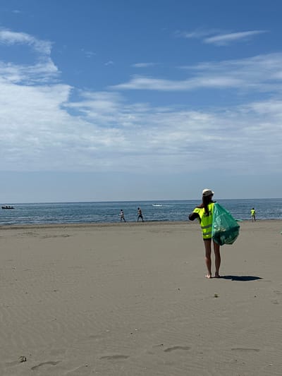 The image shows a person wearing a yellow vest and a cap standing on a sandy beach in Donji Štoj, Montenegro. The person is holding a green plastic bag. The beach is wide and flat, and the sea is visible in the background. There are other people walking along the beach in the distance. The sky is blue with some white clouds. There are some footprints on the sand.
