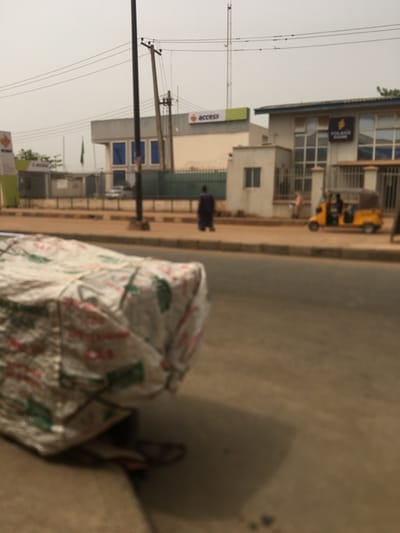 A street scene shows an Access Bank building in the background, with a yellow auto-rickshaw parked nearby. A person walks along the sidewalk. Electrical poles and wires stretch across the sky. In the foreground, a large, covered object, possibly merchandise, sits on the side of the road. The overall image is slightly blurry and tilted.