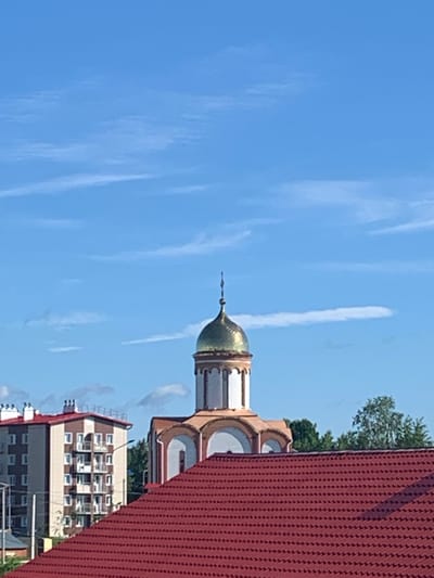 The image shows a view of the city of Volchansk, Russia, on a clear day with a blue sky and scattered clouds. In the foreground, a red-tiled roof dominates the lower portion of the frame. Behind the roof, a modern apartment building with a beige and brown facade, featuring multiple balconies and white-framed windows, stands on the left. To the right of the apartment building, a church with a distinctive golden dome and white and beige architectural details is visible. The church has arched windows and a tall, slender spire topped with a cross. Lush green trees are seen behind the church. The overall scene depicts a residential area in Volchansk with a mix of modern architecture and religious buildings under a bright sky.