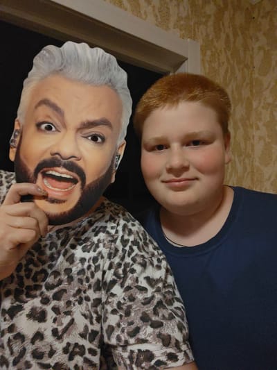 The image shows a full-body cardboard cutout of a man with gray hair and a beard, wearing a leopard-print shirt, held by a person in front of a wall with textured wallpaper. To the right of the cutout is a young person with red hair and a blue shirt. The scene appears to be indoors, possibly in a home. There is no information about the location in Markovskii, Russia.