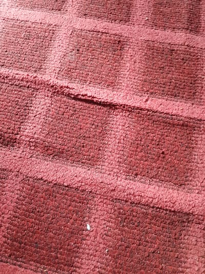 A close-up, overhead shot showcases a textured, crimson rug. The rug's surface is composed of tightly woven loops, creating a distinct ribbed pattern. The lighting highlights subtle variations in color and texture, with some areas appearing a brighter pink due to the sunlight. A few small white specks, possibly debris, are visible on the rug's surface. The image is taken in Madalla, Nigeria.