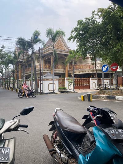 In the city of East Jakarta, Indonesia, a street scene unfolds with a large, ornate house as the backdrop. The house features a light-colored facade with elaborate gold trim on its balconies and roofline, accented by palm trees lining its front. A woman wearing a pink hijab and a patterned outfit rides a motorcycle with a child on the back down the paved street. Two motorcycles are parked in the foreground, one predominantly silver and the other a combination of black and teal. Traffic signs are visible on the right side of the road, indicating traffic direction and a prohibition. The sky is overcast, and the overall environment suggests a residential urban setting.