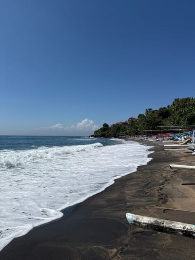 A black sand beach in Abang, Indonesia is shown under a clear blue sky. Waves with white foam crash onto the shore. Numerous traditional fishing boats, many painted with red, white, and blue accents, are lined up on the beach. Lush green vegetation covers the hillside in the background, with buildings visible among the trees. A piece of weathered wood lies in the foreground on the sand. Footprints are visible in the sand near the boats.