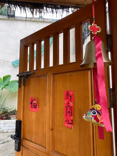 The image shows a wooden door in Dujiangyan City, China. The door is slightly ajar, revealing a glimpse of an outdoor scene. A brass bell with a red ribbon hangs from the top of the door frame. Two red paper decorations with Chinese characters are affixed to the door. One appears to say "福" (fu), meaning good fortune. The other decoration is a vertical strip with more Chinese characters and the word "HAPPY" written below. A small, colorful fabric ornament, possibly a lion's head, hangs from the bell's ribbon. A black door handle and a metal latch are visible on the door. The background shows a white wall and some greenery, suggesting an outdoor or semi-outdoor setting. The overall impression is one of a welcoming entrance decorated for a festive occasion.