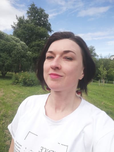 A medium shot photograph features a woman with dark hair and fair skin, looking at the camera with a slight smile. She is wearing a white t-shirt and red cherry-shaped earrings. The background is a park with green grass and trees under a blue sky with white clouds. The location is the city of Kolpino, Russia.