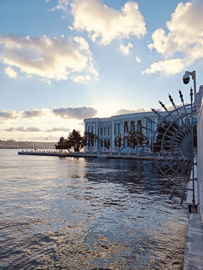 The image captures a view of the Dolmabahçe Palace in the Beşiktaş district of Istanbul, Turkey, with the Bosphorus strait in the foreground. The palace, a grand architectural structure with light-colored facades and numerous windows, is situated on the water's edge. To the right, a section of an ornate, dark-colored metal railing and part of a security camera are visible in the foreground, creating a framing effect. The water of the Bosphorus is rippled, reflecting the sky and the structures. In the middle ground, trees line the edge of the palace grounds, and across the water, a cityscape with hills and buildings is faintly visible. A Turkish flag flies on a flagpole in the distance. The sky is a mix of blue with scattered, bright white and yellowish clouds, suggesting either sunrise or sunset lighting. No people are clearly discernible in the image.