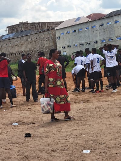 In Egbagu, Nigeria, a group of young men in white and blue soccer uniforms stand around on a dirt field. A woman in a vibrant red and green patterned dress walks past them, carrying a clear plastic bucket filled with small items. Behind the group, a multi-story building under construction with scaffolding and solar panels on its roof dominates the background. The sky is overcast with scattered clouds.