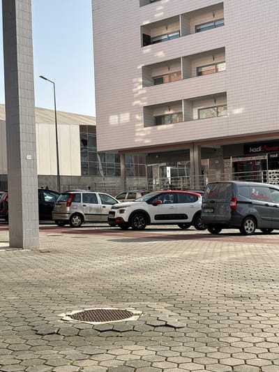 The image depicts an urban street scene in Vila Real, Portugal, lined with parked cars and adjacent to a multi-story building with a tiled facade. A gray van with a "Café Campeão" logo is parked closest to the camera, followed by a white Citroën C3 with a red roof, then several other cars. The building has a modular grid pattern on its exterior, with visible windows. A street lamp stands near a lower, horizontally-oriented structure made of similar tiles. The street is paved with interlocking hexagonal concrete blocks, and a circular manhole cover is visible in the pavement. The sky is visible in the upper left corner.