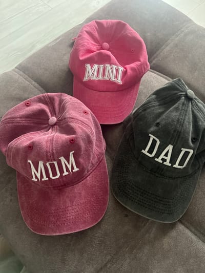 Three baseball caps are displayed on a textured, dark grey surface. The caps are arranged in a triangular formation. The cap in the top center is pink and has "MINI" embroidered in white capital letters on its front. The cap to the left is a deep pink or maroon color and has "MOM" embroidered in white capital letters. The cap to the right is dark grey and has "DAD" embroidered in white capital letters. The lighting suggests an indoor setting, possibly a couch or padded furniture. There are no people visible in the image. The image does not provide any information about its location in Chelyabinsk, Russia.