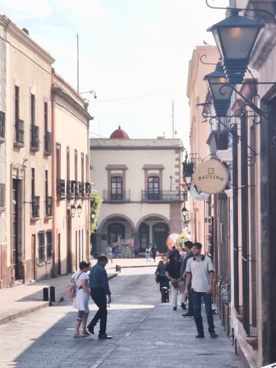 This street scene is in the city of Querétaro, Mexico, with cobblestone streets lined by pale pink buildings. Three old-fashioned street lamps are on the right, affixed to the buildings with decorative ironwork. One sign reads "Paulino." In the foreground, a woman in a white dress and pink backpack walks with a man in a blue plaid shirt. Further down the street, a group of people is walking, including a man in a striped shirt. In the distance, an arched building with a red dome is visible. People are milling about in that area.
