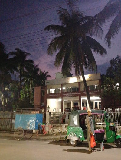 The image depicts a street scene in Bhola, Bangladesh, located at the coordinates 22.657 latitude and 90.663 longitude. It is dusk, and a single man is standing by a green auto rickshaw. The rickshaw is parked in front of a two-story building, possibly an office or government building. The building has a sign that reads "Bangladesh Bank Bhola Branch." The man is dressed in a long white garment and a cap. He is holding a red bag and looking at the camera. There is a bicycle parked next to the rickshaw, and a blue and white cart is parked further down the street. The street is paved and lined with trees, including palm trees and other foliage. Overhead power lines are visible. The sky is a dark purple color.