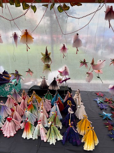 The image depicts a display of numerous handmade fairy figurines, crafted from fabric and other materials, arranged on a black surface. The figurines vary in color and design, many featuring voluminous, layered skirts and delicate, articulated limbs. Some fairies are suspended from a netting background by thin threads, while others stand upright on the surface. The background consists of a light-colored netting with a blurred outdoor scene visible through it, possibly suggesting a market stall or outdoor exhibition. Decorative beads and faux leaves are strung across the top of the netting. The overall impression is a whimsical collection of artistic creations, possibly for sale or exhibition. The location is identified as Vracar Urban Municipality, Serbia.