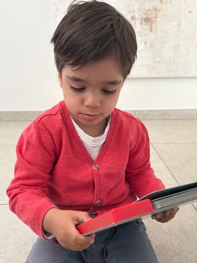 A high-angle, portrait-oriented shot, rotated 90 degrees clockwise, captures a young child intently focused on a book indoors in Junglinster, Luxembourg. The child, with dark hair and long eyelashes, is shown from the chest up, positioned on a light-colored, possibly tiled floor. Their eyes are cast downwards, suggesting deep concentration or peaceful engagement with the book.
The child is wearing a vibrant red knitted cardigan with brown buttons over a white t-shirt, and blue-grey pants are visible from the waist down. Their hands gently hold an open book, which has a distinct red spine or cover. The pages of the book are visible between their hands.
The background consists of a plain light-colored wall or surface, possibly with some subtle texture, creating a minimalist setting that keeps the focus on the child and the book. The lighting is bright and even, indicating daytime. No specific time of day or weather can be discerned. There is no visible text in the image. The overall impression is one of quiet learning or imaginative play.