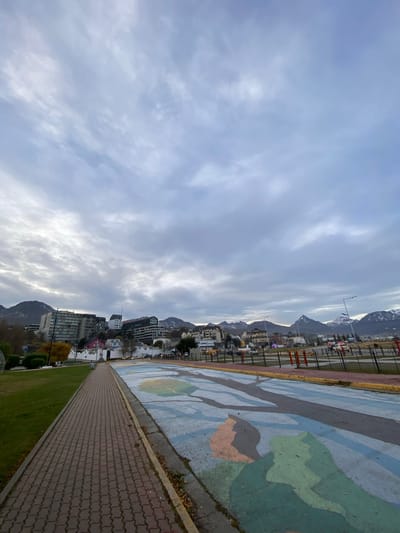 The image depicts a cityscape of Ushuaia, Argentina, seen under a cloudy sky. The cityscape is oriented horizontally, with buildings of varying heights clustered along the edge of mountains. A paved walkway, with a brick pattern, runs along the right side of the frame, adjacent to a grassy area and a section painted with abstract shapes. In the background, snow-capped mountains are visible. The image's perspective suggests it was taken from an elevated position, possibly along a waterfront or a park area that offers views of the city and the surrounding landscape.