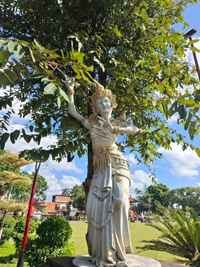 A full-shot, eye-level, outdoor, medium-angle, natural light photograph captures a stone statue of a Balinese dancer situated in front of a large tree with green foliage and a blue sky with scattered clouds. The statue, primarily white with gold accents, depicts a woman in traditional Balinese attire, including an elaborate headdress, ornate jewelry, and flowing garments. Her arms are raised, and her hands are positioned in a dance-like gesture. The statue is positioned on a stone pedestal. Behind the statue, a lush green lawn stretches towards a cluster of buildings with orange tiled roofs. Several trees, including palm trees, are visible in the background, along with a red pole with a black fixture. The overall scene suggests an outdoor public space, possibly a park or a cultural site, in Gianyar, Indonesia.