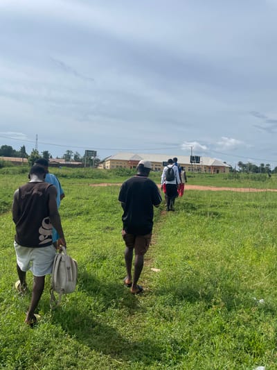 The image captures a group of people walking in a line across a sunlit green grass field in Ogbogbo, Nigeria. The perspective is from behind the group, with individuals moving away from the viewer along a dirt path that traverses the field.
In the foreground, a person is visible from the back, wearing a brown t-shirt with the text "Summer Society" printed on it, light-colored shorts, and carrying a beige backpack. Ahead of them, another individual wears a black t-shirt, dark shorts, and a white baseball cap. Further down the line, several more people are seen, some wearing white shirts, one in blue, and another in red.
The setting features a wide expanse of green grass. In the mid-ground, a metal-framed sign or board stands near the path. The background reveals light-colored buildings, possibly institutional or residential, with distinct roofs, under a clear, bright blue sky with scattered white clouds. Sparse trees and utility lines are also visible in the distance.
The scene is well-lit by natural daylight, suggesting it is mid-morning or early afternoon, with clear shadows cast by the people and structures. The overall impression is of a group undertaking a walk or procession through an open, possibly campus or community, area.