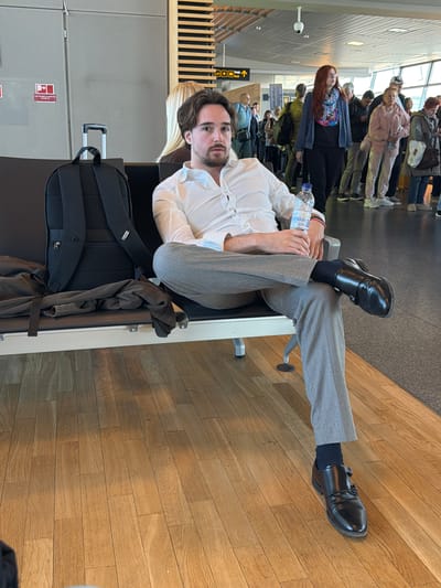 The image captures an indoor scene, likely a waiting area or departure lounge at Rīga International Airport in Latvia. The main subject is a man, appearing to be in his late 20s or 30s, with dark hair and a beard, wearing a white long-sleeved shirt and light grey trousers. He is seated on a bench, looking directly at the camera with a neutral expression, and holds a clear plastic water bottle with his hands. His left leg is crossed over his right, showcasing black double monk strap shoes and dark socks.
Next to him, a black backpack with a prominent handle, typical of carry-on luggage, rests on the floor, accompanied by a dark jacket draped over it or the seat. The floor is made of light-colored wooden planks.
In the background, a line of people is visible, suggesting a queue or waiting area. Among them, a woman with long red hair and a patterned top stands out. Overhead, white ceiling panels with integrated lighting are visible. A yellow sign with a left arrow and suitcase icon is present in the mid-background, indicating directions, possibly for baggage claim or check-in. Other red signs, too small to be legible, are also visible on a grey wall. The overall ambiance is that of a bustling but organized public transport hub. The scene appears to be taking place during daylight hours, given the bright interior lighting.