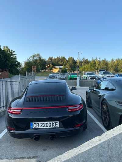 The image shows a parking lot in Kerenite, Bulgaria. In the foreground, two black Porsche 911s are parked side-by-side. The car on the left has its rear visible, displaying a license plate that reads "CB 2200 KE" and "Porsche Zentrum Hagen" below it. The car on the right is also a Porsche 911, dark green in color, with its side and rear wheel visible. In the background, several other cars are parked, including a bright green Porsche. The parking lot is surrounded by trees and a clear blue sky. A metal fence is visible to the left of the black Porsche, and a sign with a "P" symbol for parking is mounted on it. Buildings with wooden structures are also visible in the background, suggesting a resort or rural area. The lighting indicates it is either early morning or late afternoon.
