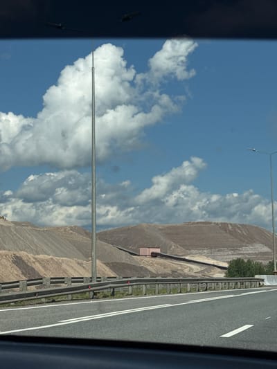 A wide-angle, eye-level, exterior shot depicts a view from a moving vehicle on a paved highway. The highway, with its white lane markings, occupies the foreground, with a metal guardrail to the left. Beyond the guardrail, large mounds of pale brown material, likely industrial waste or spoils, dominate the landscape. A conveyor belt system is visible ascending one of the mounds. In the background, a bright blue sky is filled with fluffy white clouds. Two tall, silver lampposts are positioned in the frame, one closer and to the left, the other further away and to the right. A pink industrial building is visible nestled near the base of a mound. The scene is characteristic of the Solikamsk potassium mining region in Russia. There are no people or animals visible in the image.