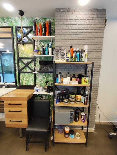 This image shows a barbershop or hair salon located in the city of Sarıyer, Turkey. The scene is composed of a wooden desk with drawers on the left, a black chair in front of the desk, and a multi-tiered shelf unit filled with hair products and tools against a brick-patterned wall. The background features a wall with a tropical leaf pattern.
The wooden desk has a light wood finish with three drawers, each with a black handle. The top of the desk has a white sink on the left side. A black leather chair with a low back is positioned in front of the desk.
The shelf unit is made of black metal with wooden shelves. The top shelf holds several cans of hairspray, mostly in red, orange, and black packaging, and some smaller containers of hair products. The second shelf contains various bottles of hair products, including shampoos, conditioners, and styling sprays, as well as some white cotton pads. A black case holding several pairs of scissors is also on this shelf. The third shelf has a grey appliance, likely a sterilizer or warmer, a black spray bottle, a bottle labeled "Baleaf", and a purple box. The bottom shelf displays a variety of hair waxes, pomades, and gels in different colored containers.
The wall behind the desk and shelving unit is covered in wallpaper with a vibrant green tropical leaf pattern. To the right of the shelving unit, the wall has a textured grey finish that mimics brickwork. A black pendant light with a rope cord hangs from the ceiling. The floor is a dark, smooth surface, possibly concrete or polished wood.
There are no people visible in the image. The overall impression is that of a well-organized and equipped space for hair care services.