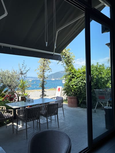View from an open doorway of an outdoor dining area in Tivat, Montenegro. The scene captures a table and chairs arranged on a patio, with a partial view of a seated person. Outside, there is a view of a beach, trees, and a body of water with boats. A mountain is visible in the distance. A retracted awning partially covers the view from the left, and greenery is present in planters and surrounding areas. A shopping cart is seen near a building exterior behind the planter. The sky is clear and blue.
