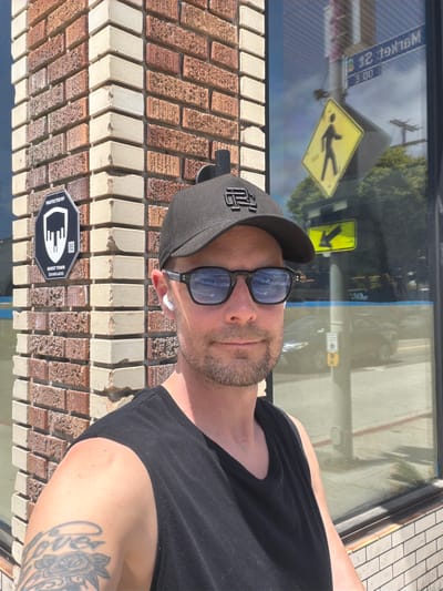 A man wearing a black tank top, a black baseball cap, and sunglasses takes a selfie outdoors. He is standing in front of a brick building with a window reflecting a street sign that reads "MARKET ST 13 00". A pedestrian crossing sign is also visible in the reflection. He has a beard and a tattoo on his left arm. He is wearing white wireless earbuds.