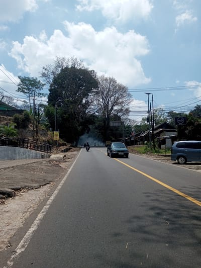 The image depicts a straight, asphalt road in a semi-urban setting. A yellow dividing line runs down the center of the road. A dark green car is driving on the right side of the road, and two motorcyclists are further down the road. On the left side of the road is a raised, broken sidewalk with vegetation growing on the embankment. On the right side of the road, there is a light blue van parked next to a small building with a sign in Arabic. Overhead power lines cross the sky. The sky is light blue with scattered white clouds. There is sparse smoke visible behind the large trees in the background. The location appears to be outside the city center, possibly in a rural area of Nawng Hkar, Myanmar.