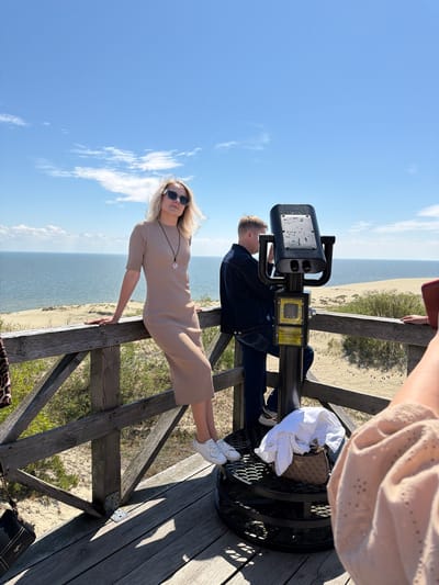 The image captures a scene on a wooden observation deck overlooking a sandy landscape and the sea under a clear blue sky with scattered clouds. In the foreground, a woman with blonde hair and sunglasses is leaning on a wooden railing, wearing a beige knit dress and white sneakers. To her right, a man in a denim jacket and jeans is looking through a large telescope. A small, patterned handbag and a white shirt are placed on the base of the telescope. In the far right, a portion of another person's arm and shoulder, wearing a light-colored, textured garment, is visible. The environment suggests a coastal or dune area, possibly within the vicinity of Morskoye, Russia, known for its sand dunes.