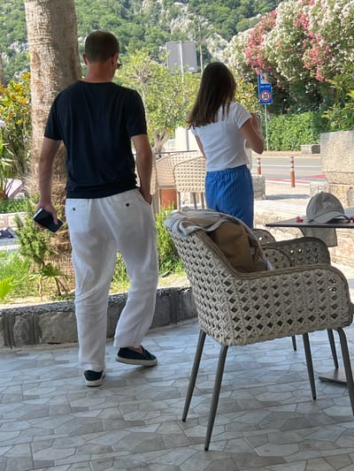A man wearing a black t-shirt and white linen pants walks away from the camera, holding a smartphone in his left hand. Next to him, a woman in a white t-shirt and blue striped pants stands looking away from the camera. The scene appears to be outdoors, possibly a cafe or restaurant patio, with wicker chairs and a table visible. A sign with "30" in a red circle is visible on a post in the background, suggesting a speed limit. The background shows lush greenery and a mountainous landscape, consistent with the town of Risan, Montenegro.