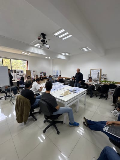 The image shows an indoor scene, apparently a classroom or studio, in San Andrés Cholula, Mexico. There are approximately 20-25 people present, most of whom appear to be students. They are gathered around several white tables on which architectural models and drawings are displayed. An older man, possibly an instructor, stands at one of the tables and appears to be lecturing or giving feedback. Most of the students are seated in chairs, listening or observing. Some are leaning forward, engaged with the presentation. In the background, there are posters and architectural drawings displayed on the walls. The room has white walls, tiled floors, and large windows that let in natural light. A projector is mounted on the ceiling. The atmosphere seems to be one of a design review or critique session.
