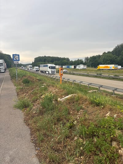 The image depicts a traffic jam on a highway, likely in or near Heidenheim an der Brenz, Germany. A line of trucks and cars are stopped or moving slowly in multiple lanes. On the side of the road, a pedestrian walkway is visible, with an SOS sign indicating an emergency phone. Vegetation and trees line the highway and beyond, suggesting a rural or semi-rural setting. The sky is overcast. The scene appears to be a typical highway scene, possibly indicating a temporary traffic delay.
