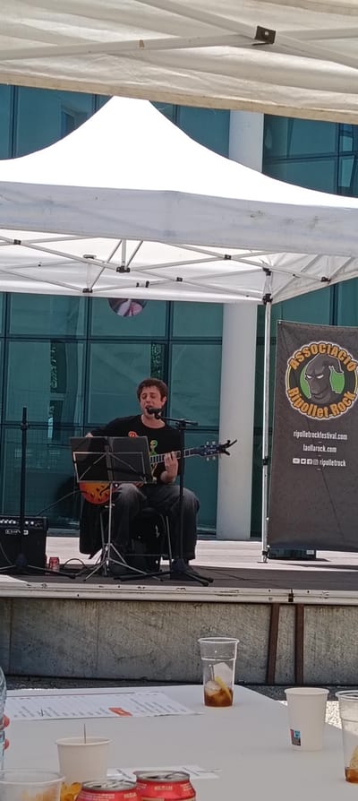 The image shows a man performing on a small stage outdoors, under a white canopy. He is sitting on a chair, playing an electric guitar, and singing into a microphone. He's wearing a black t-shirt with a graphic on it and jeans. Next to him is an amplifier and a music stand. Behind the stage is a banner for "Associació Ripollet Rock" with a logo featuring a hooded figure and website addresses. The backdrop behind the stage is a modern building with glass windows. In the foreground, there's a table with plastic cups, a soda can, and some other items. The event appears to be taking place in the city of Ripollet, Spain based on the banner.