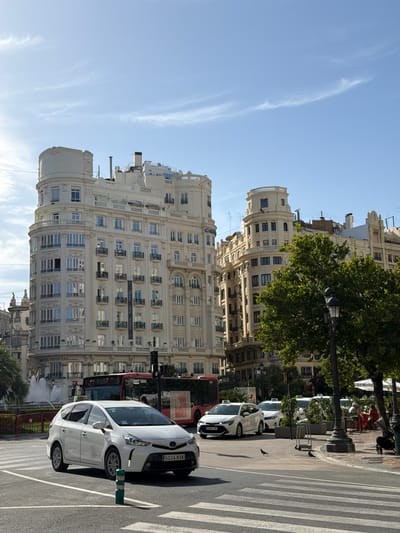 The image captures a street scene in the city of València, Spain, likely in the vicinity of Plaça de l'Ajuntament, given the architectural style and presence of a bus stop with signage that could be associated with public transport hubs in the city. In the foreground, a white Toyota Prius taxi, license plate 5024 XKX, is turning at an intersection with a pedestrian crossing. Another white taxi is partially visible to the right of the Prius. Behind the taxis, a red bus is stopped, possibly at a bus stop, with people visible inside.
Dominating the background are several large, ornate white buildings with multiple stories, featuring numerous windows, balconies with wrought-iron railings, and decorative architectural elements. One building prominently displays signage that reads "HOTEL VENECIA" and "VALENCIA PLAZA CENTRO," confirming the location. A fountain is visible to the far left, with water jets spraying upwards. Green trees line the right side of the street, with tables and chairs set up at an outdoor cafe area beneath them, where a few people are seated. The sky is blue with scattered light clouds, suggesting a sunny day. The overall impression is of a lively urban square in a European city.