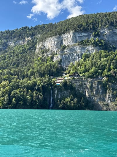 The image depicts a stunning view of a turquoise lake, likely Lake Thun in Switzerland, with a dramatic cliff face and lush green forest rising behind it. Several buildings are nestled on ledges along the cliffside, including a prominent structure with a dark roof. A waterfall cascades down the rocky face, adding to the picturesque scenery. The sky above is a vibrant blue with scattered white clouds. The water is choppy, suggesting a gentle breeze. No people or animals are visible in the frame.