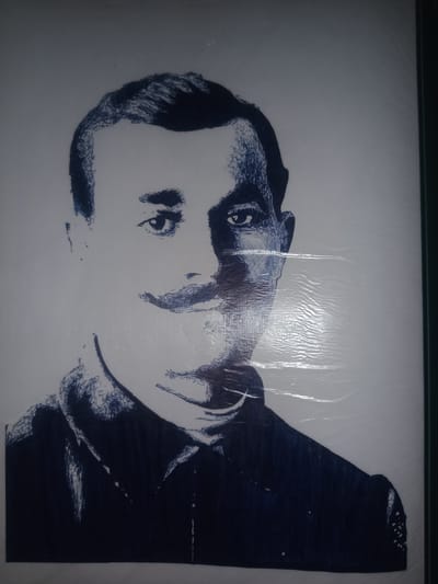 This is a close-up, full-body stencil portrait of a man. The portrait is rendered in dark blue against a white background, with varying densities of blue indicating shading and detail. The man is depicted from the chest up, facing slightly to his left, with his gaze directed forward. He has a prominent mustache and appears to be wearing a collared shirt or uniform with several visible buttons. The style is illustrative and somewhat abstract, with energetic lines used to create texture and depth, particularly in his hair and the folds of his clothing. The image appears to be a photograph of a physical artwork, possibly a poster or a mural, as there are visible horizontal lines across the surface, suggesting it might be covered with plastic sheeting or is on a textured surface. The lighting in the photograph creates some glare and reflections on the surface. The location context provided is Gulbag, Uzbekistan.