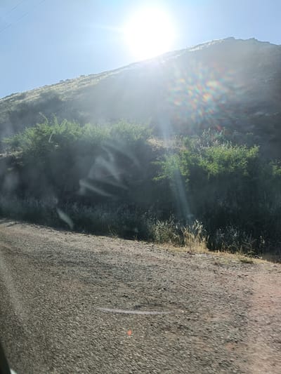 The image shows a hillside landscape in Bin El Ouidane, Morocco. The sun is high in the sky, creating a bright glare and lens flare. A dirt or gravel road is in the foreground. The hillside is covered in vegetation, including bushes and grasses. The light is very bright which may obscure some details.
