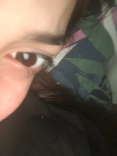 A close-up shot focuses on a brown eye with light reflecting in the pupil. The eyebrow is visible above, and the side of the nose is in the foreground. In the background, there's a bed sheet with a pattern of green, pink, and blue shapes. To the right of the eye, dark hair and a black surface dominate, creating a contrast. The overall composition is intimate and somewhat obscured.