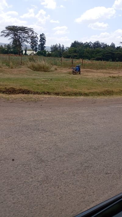 An asphalt road is in the foreground, with a strip of green grass bordering it. Beyond the grass, a field stretches out with a parked motorbike on it. A fence runs along the field's edge, separating it from a line of trees. In the distance, a few buildings are visible, set against a backdrop of rolling hills. The sky is bright with scattered clouds.