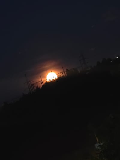 A partially eclipsed sun, appearing as a bright orange crescent with a white core, is setting behind a silhouetted treeline and a power line infrastructure in Malaya Topka, Russia. The sky is dark blue with hints of pink and orange near the horizon, suggesting dusk. Tall, skeletal power pylons and transmission lines are visible against the sky and the dark landscape. The foreground is dominated by the dark mass of trees and terrain, with some foliage visible in the lower right portion of the image. The image is taken at a downward angle, with the horizon tilted. There are no people or animals present in the frame. The overall scene depicts a natural phenomenon occurring within an industrialized landscape.